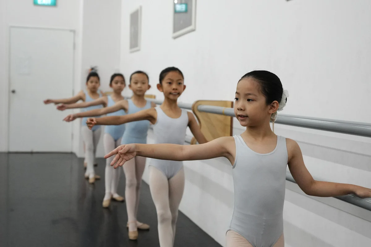 Ballet students in pale blue leotards and tights learning together at a studio barre.