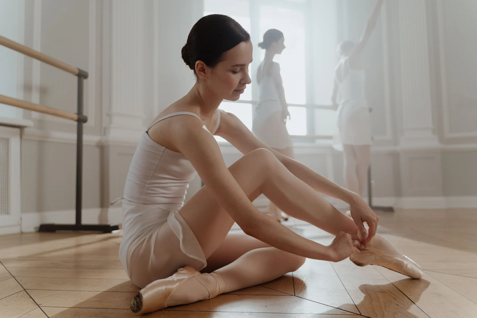 A dancer with dark hair in a ballet bun seated in a studio tying her pointe shoes
