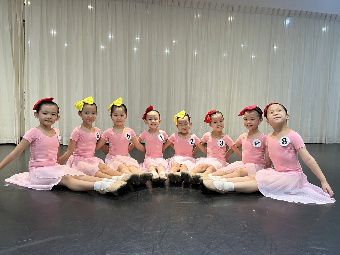 Eight young ballet students seated together in pink leotards and skirts, with colourful hair bows and ballet slippers.